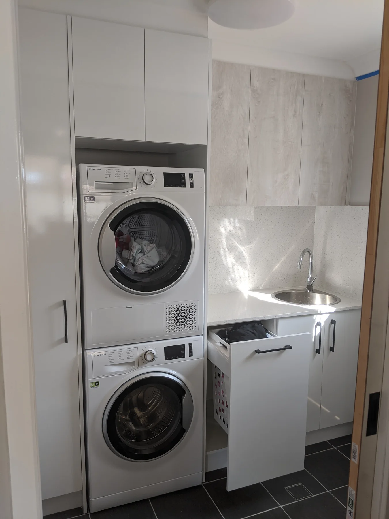 Compact completed laundry with stacked appliances, white cabinetry, a round sink and pull-out hamper